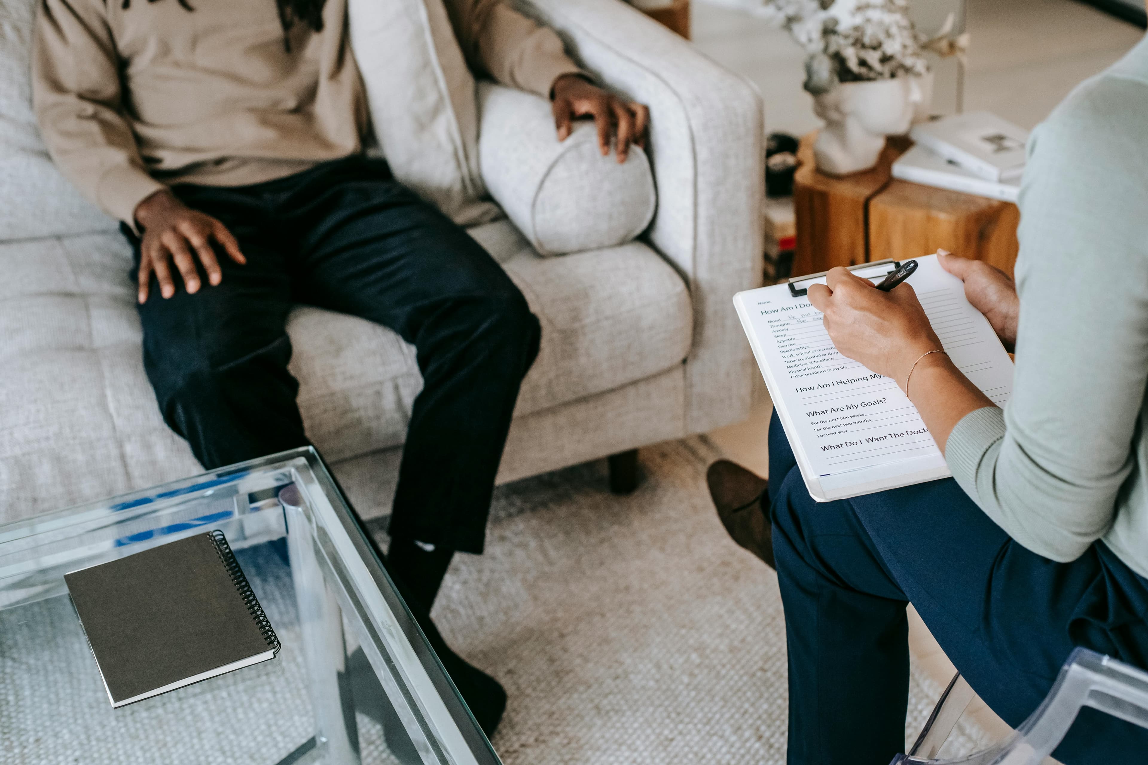 Diagnostische Situation: eine Fachperson macht mit Stift Notizen auf einem Anamnese-Fragebogen, während eine Person auf dem Sofa sitzt