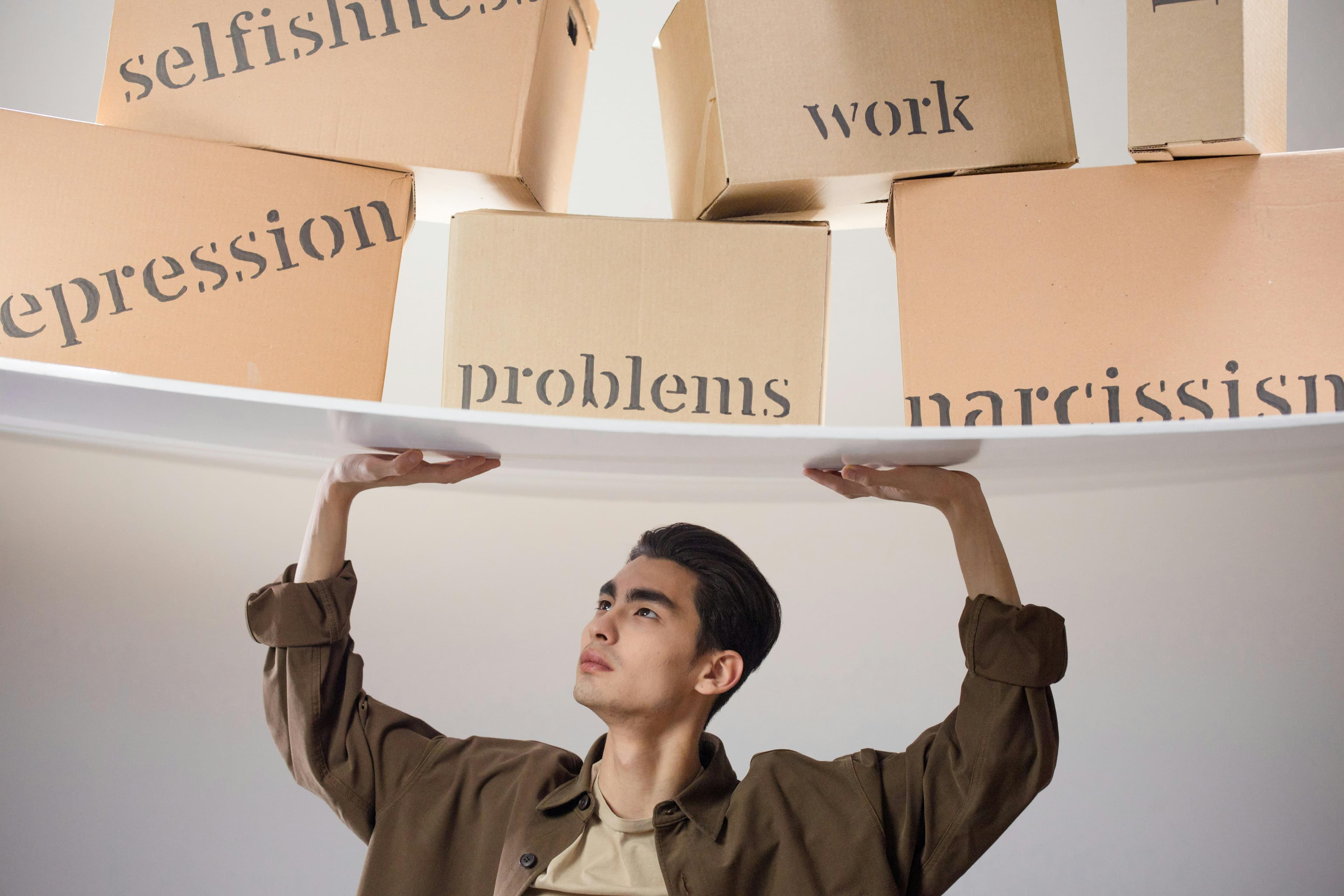 Ein junger Mann stemmt über dem Kopf eine Ablage voller Kartons, beschriftet mit ‚selfishness‘, ‚depression‘, ‚problems‘ und ‚narcissism‘