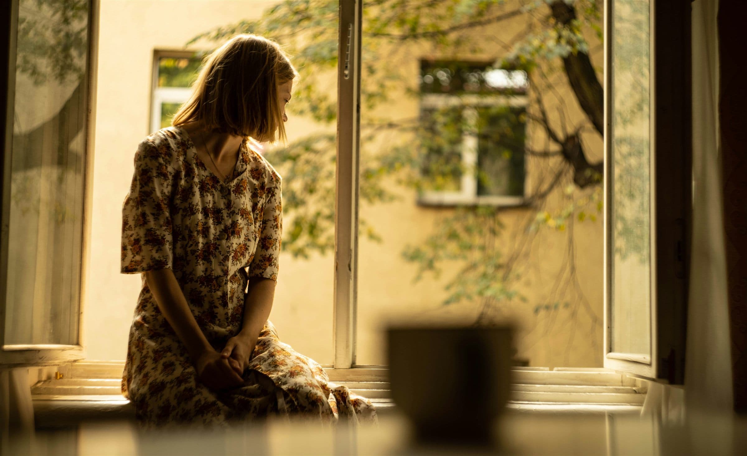 Eine junge Frau im geblümten Kleid sitzt nachdenklich am offenen Fenster im warmen Abendlicht und blickt still nach unten – Sinnbild für das lange Übersehenwerden von Frauen mit AuDHS.